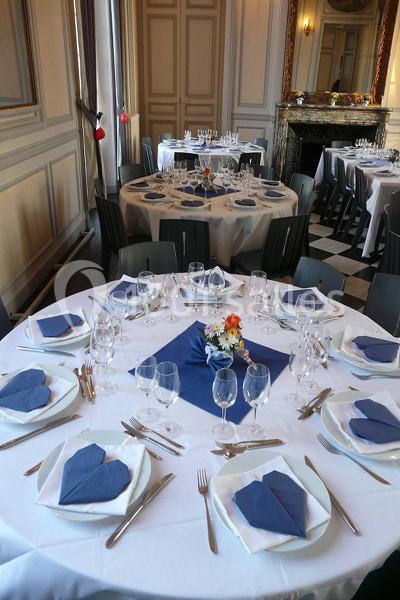 Salle de réception avec tables rondes dressées, nappes blanches, serviettes bleues pliées et décoration florale.