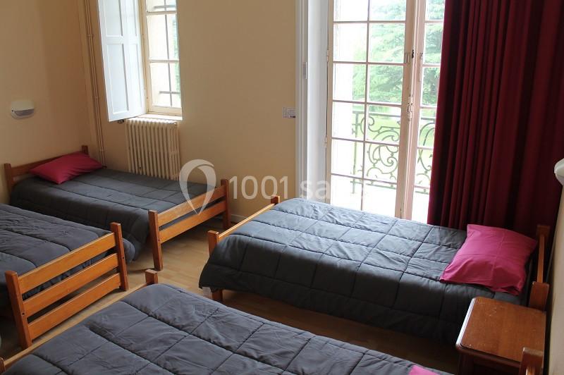 Chambre avec trois lits simples, draps gris, oreillers roses, fenêtre ouverte donnant sur un balcon et vue verdoyante.