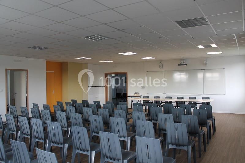 Salle de conférence vide avec rangées de chaises grises, tableau blanc et projecteur au mur.