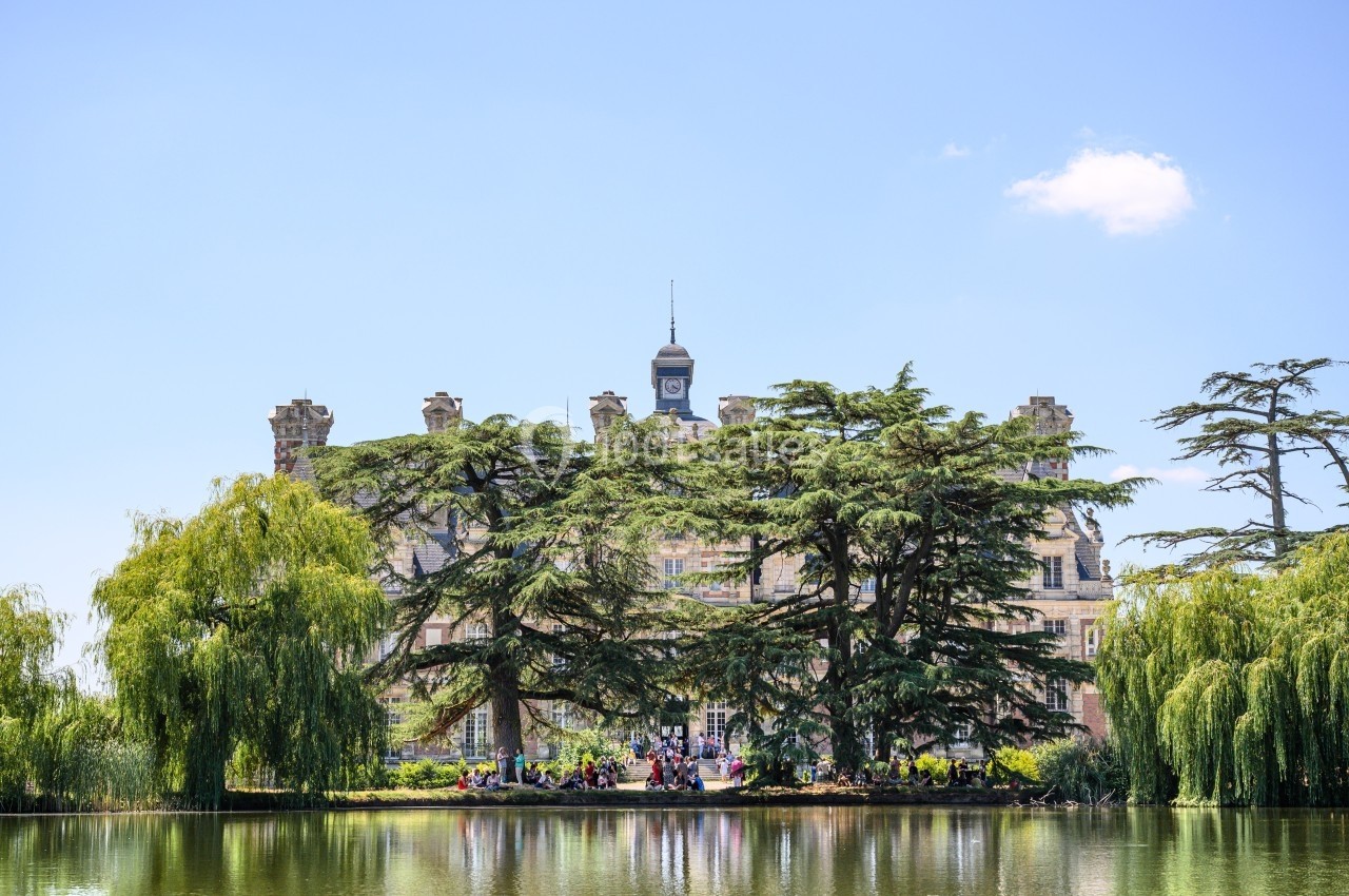 Façade d'un château entouré d'arbres, avec un plan d'eau au premier plan sous un ciel dégagé.