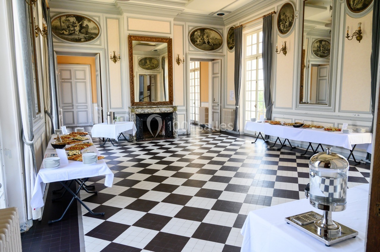 Salle élégante avec sol en damier, tables dressées pour un buffet et grandes fenêtres laissant entrer la lumière naturelle.