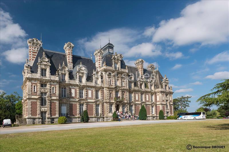 Façade d'un château en pierre et brique avec des toits en ardoise, entouré d'une pelouse et d'arbres sous un ciel bleu.