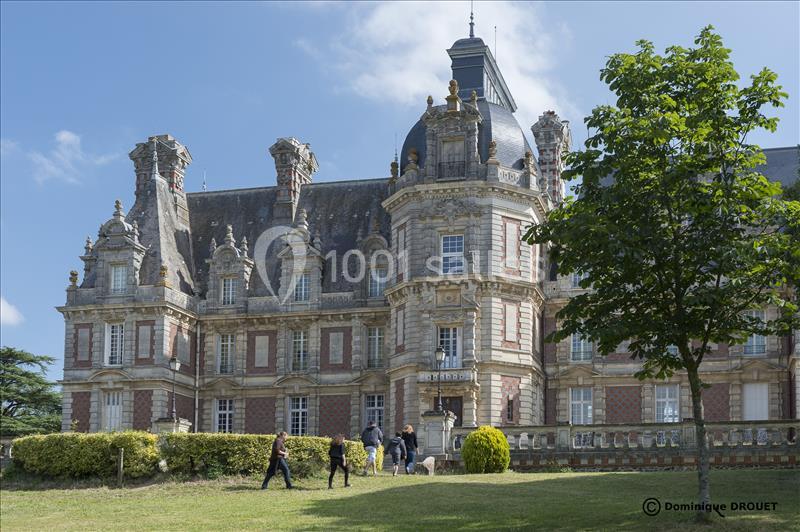 Façade d'un château en pierre et briques rouges avec des visiteurs dans un parc verdoyant par temps ensoleillé.