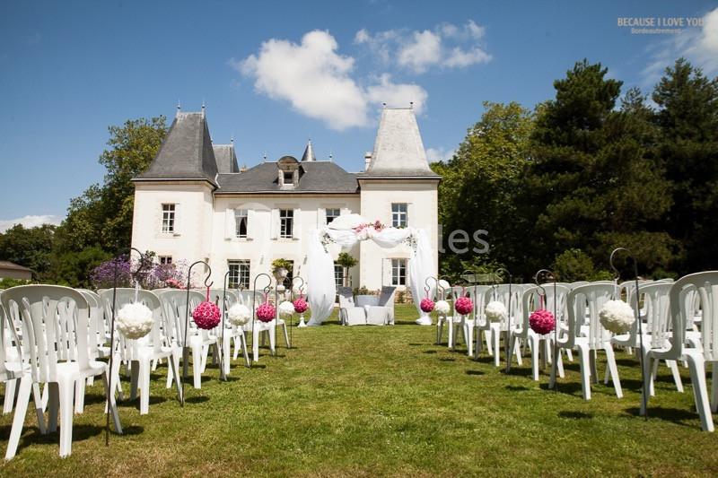 Allée de chaises blanches décorées de fleurs roses et blanches, menant à une arche de mariage devant un château.