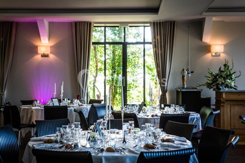 Salle de réception élégante avec tables rondes dressées, décor floral et lumière naturelle entrant par de grandes fenêtres.