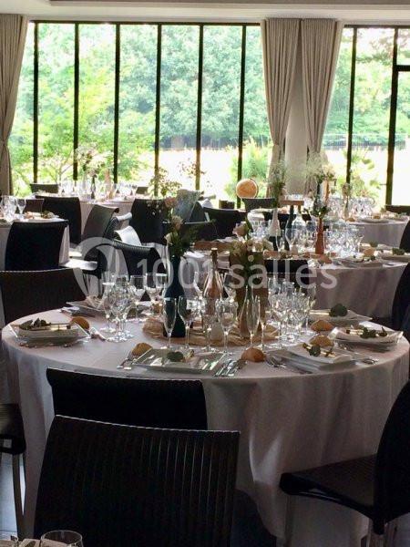 Salle de réception lumineuse avec tables rondes dressées, nappes blanches, vaisselle élégante et vue sur un jardin.