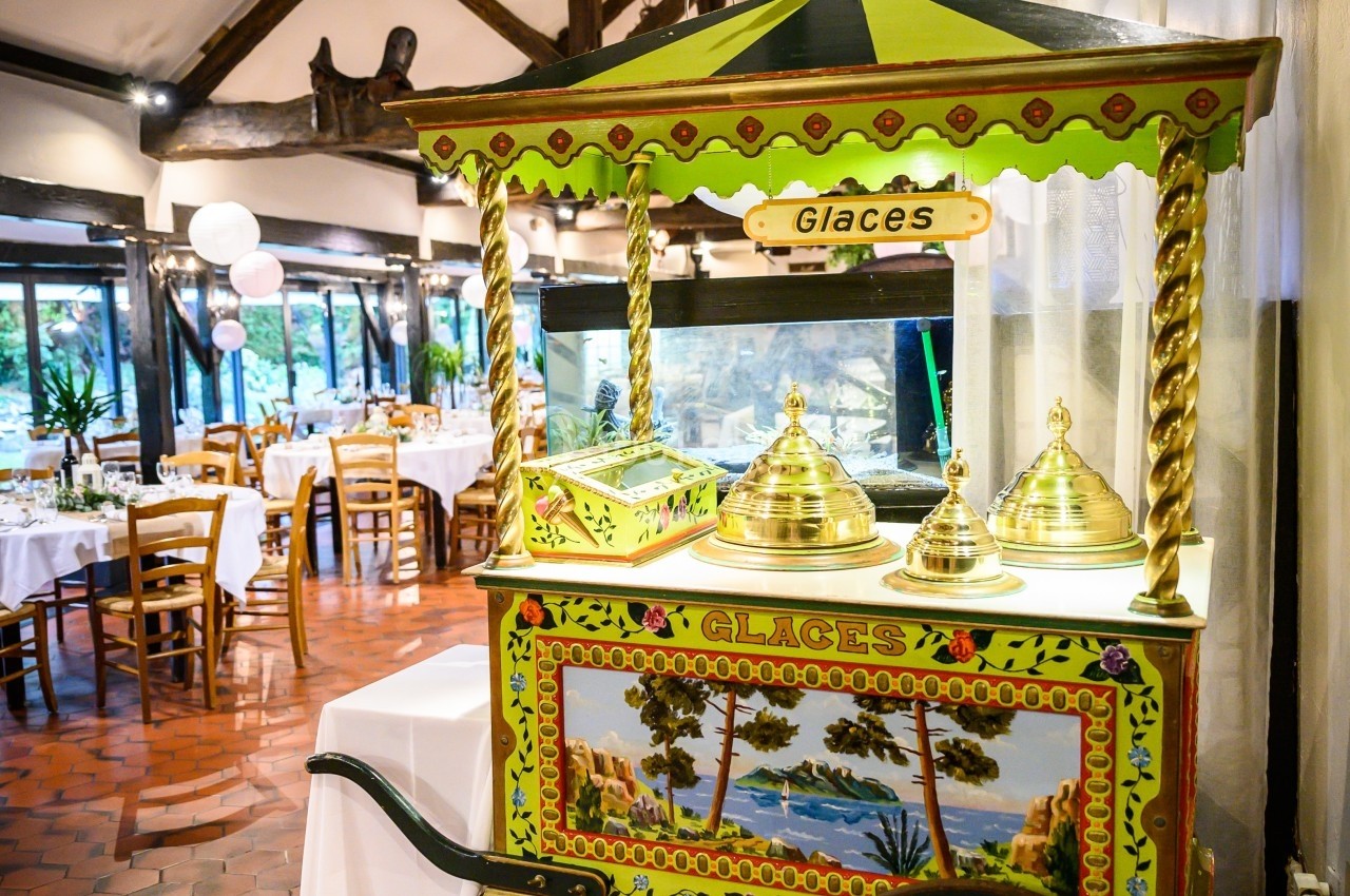 Chariot de glaces coloré avec couvercles dorés, installé dans une salle de réception au sol en carreaux rouges.