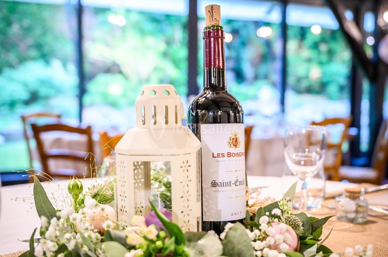 Bouteille de vin rouge posée sur une table décorée de feuillage, fleurs blanches et une lanterne blanche.