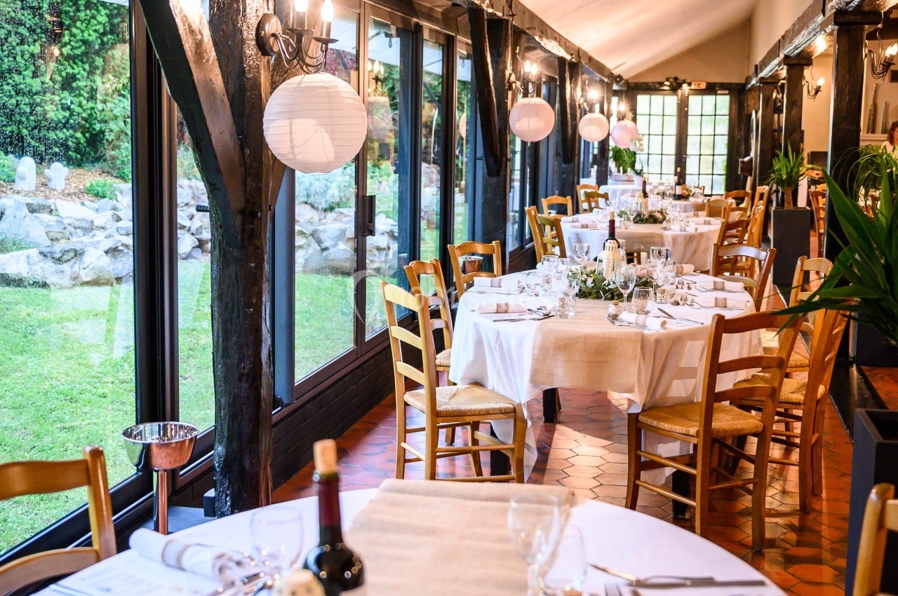 Salle de réception lumineuse avec tables décorées, nappes blanches, chaises en bois et vue sur un jardin extérieur.