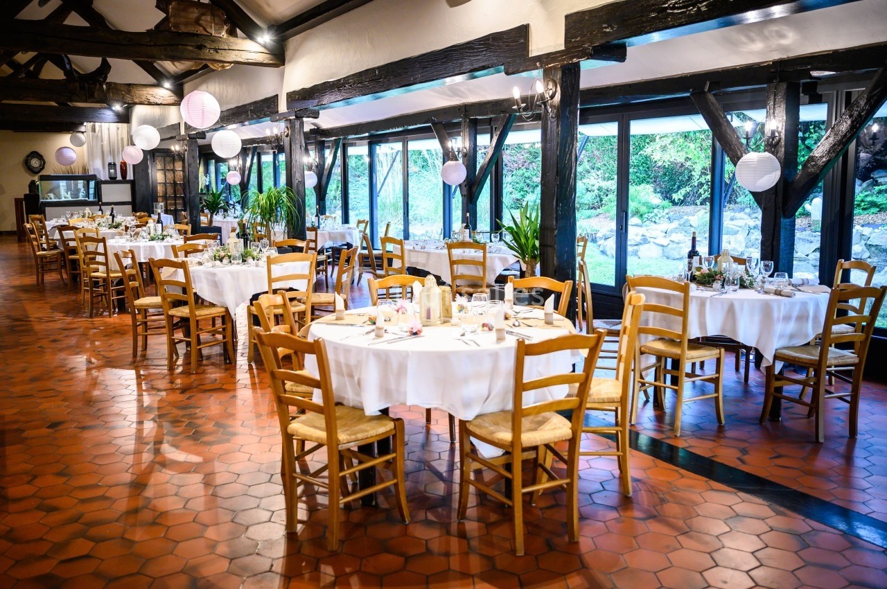 Salle de réception lumineuse avec tables rondes dressées, chaises en bois et grandes baies vitrées donnant sur un jardin.