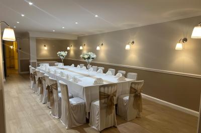Salle lumineuse avec une grande table rectangulaire dressée, entourée de chaises décorées, parquet clair et fenêtres donnant…