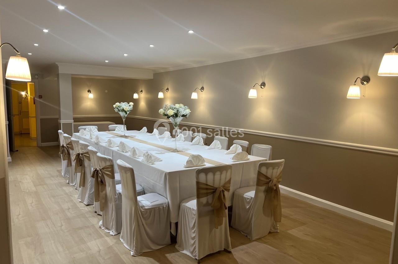 Salle élégante avec deux longues tables dressées, nappes blanches, chaises décorées et éclairage chaleureux.