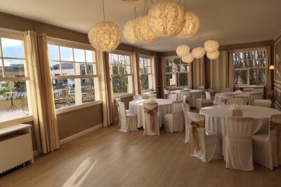 Salle lumineuse avec une grande table rectangulaire dressée, entourée de chaises décorées, parquet clair et fenêtres donnant…