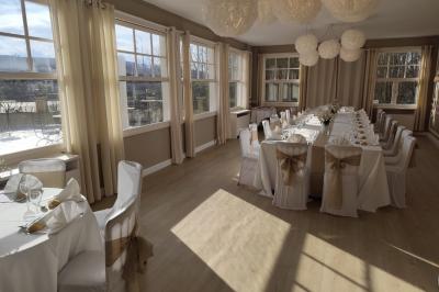 Salle lumineuse avec une grande table rectangulaire dressée, entourée de chaises décorées, parquet clair et fenêtres donnant…