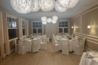 Salle lumineuse avec une grande table rectangulaire dressée, entourée de chaises décorées, parquet clair et fenêtres donnant…