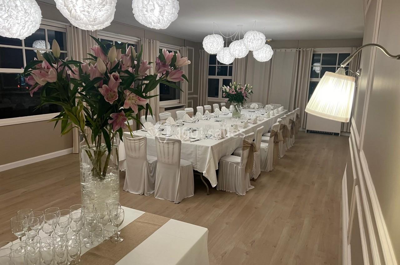 Salle de réception élégante avec tables dressées, chaises blanches et décorations lumineuses suspendues.
