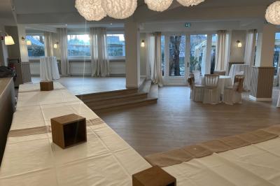 Salle lumineuse avec une grande table rectangulaire dressée, entourée de chaises décorées, parquet clair et fenêtres donnant…