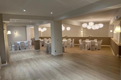 Salle lumineuse avec une grande table rectangulaire dressée, entourée de chaises décorées, parquet clair et fenêtres donnant…