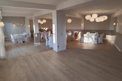 Salle lumineuse avec une grande table rectangulaire dressée, entourée de chaises décorées, parquet clair et fenêtres donnant…