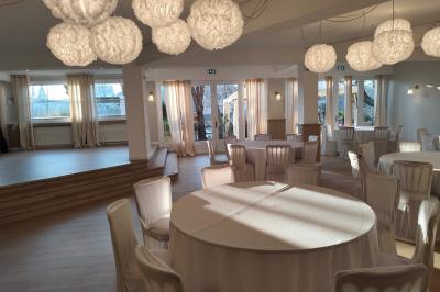 Salle lumineuse avec une grande table rectangulaire dressée, entourée de chaises décorées, parquet clair et fenêtres donnant…