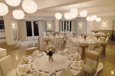 Salle lumineuse avec une grande table rectangulaire dressée, entourée de chaises décorées, parquet clair et fenêtres donnant…