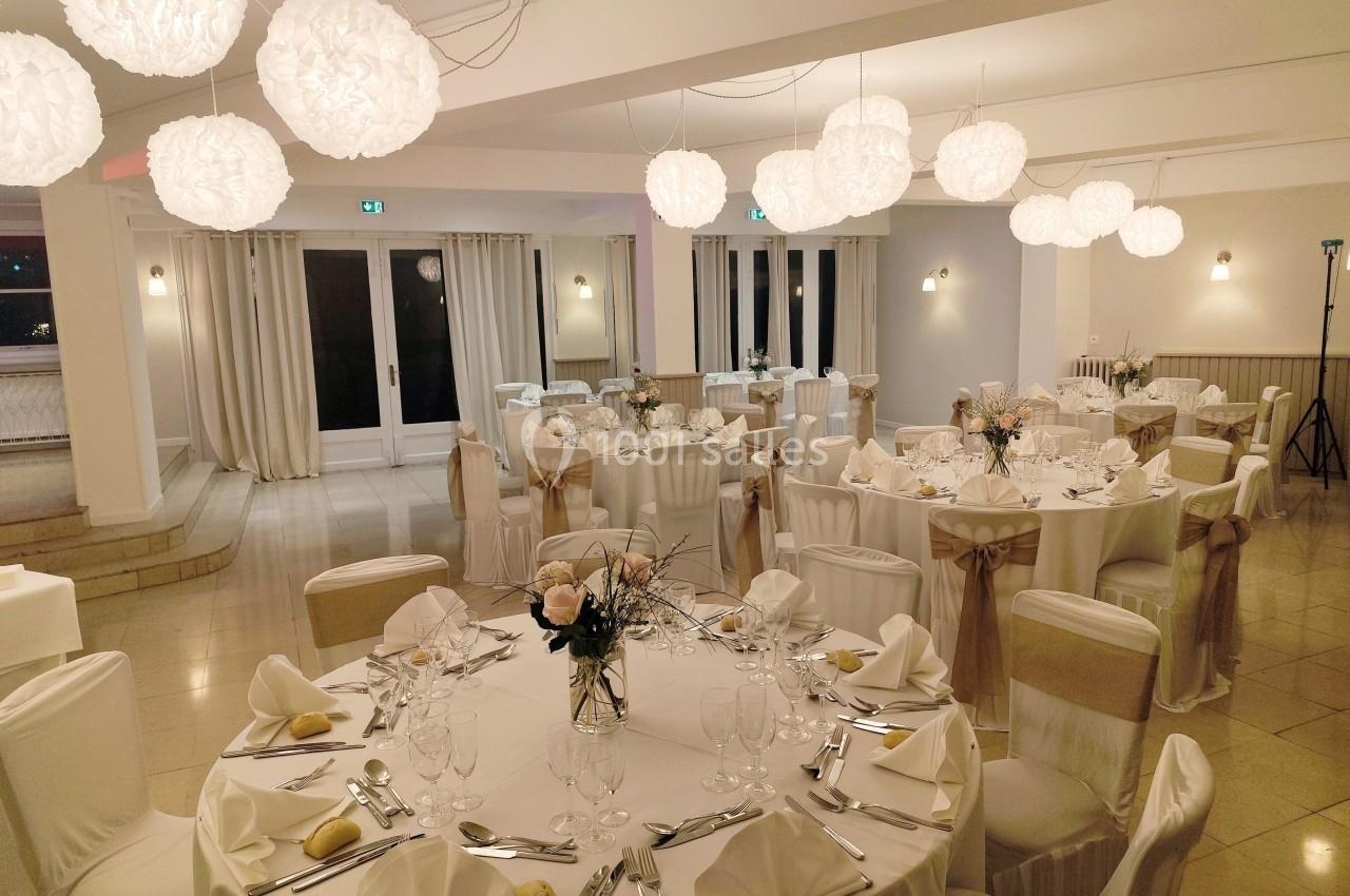 Salle de réception élégamment décorée avec des tables rondes dressées, des chaises habillées et des luminaires suspendus.