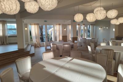 Salle lumineuse avec une grande table rectangulaire dressée, entourée de chaises décorées, parquet clair et fenêtres donnant…