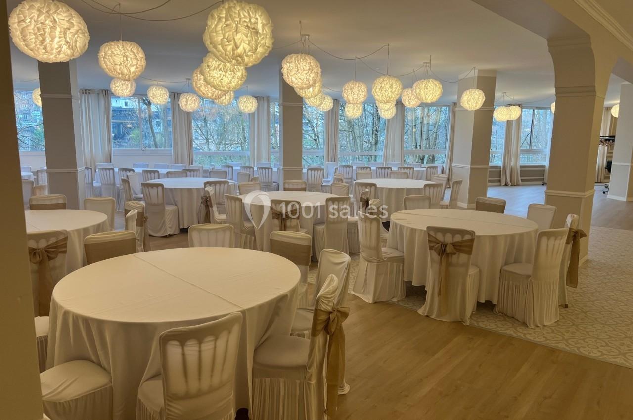 Salle de réception lumineuse avec des tables rondes et des chaises décorées de housses blanches et nœuds beiges.