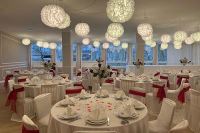 Salle lumineuse avec une grande table rectangulaire dressée, entourée de chaises décorées, parquet clair et fenêtres donnant…