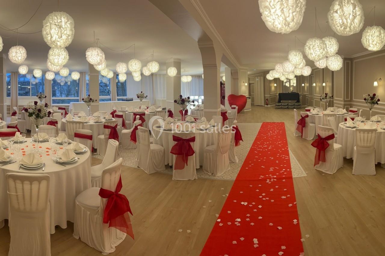 Salle de réception décorée avec des tables rondes, nappes blanches, nœuds rouges et un tapis rouge central.