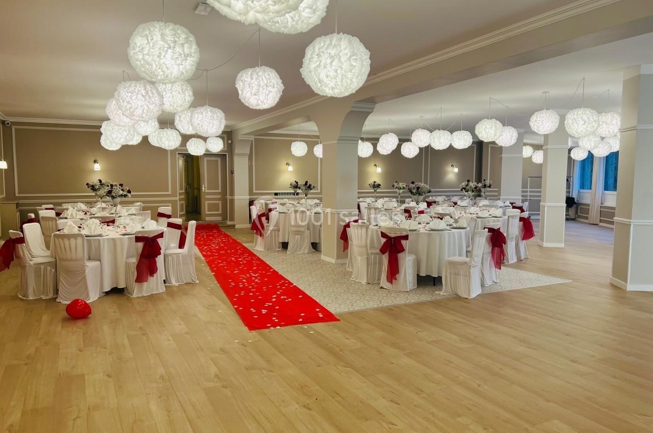 Salle de réception décorée avec des tables rondes, nappes blanches, nœuds rouges et un tapis rouge au centre.