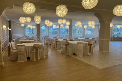 Salle lumineuse avec une grande table rectangulaire dressée, entourée de chaises décorées, parquet clair et fenêtres donnant…