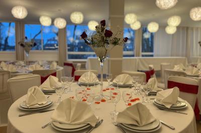 Salle lumineuse avec une grande table rectangulaire dressée, entourée de chaises décorées, parquet clair et fenêtres donnant…