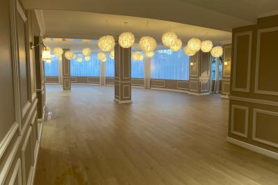 Salle lumineuse avec une grande table rectangulaire dressée, entourée de chaises décorées, parquet clair et fenêtres donnant…