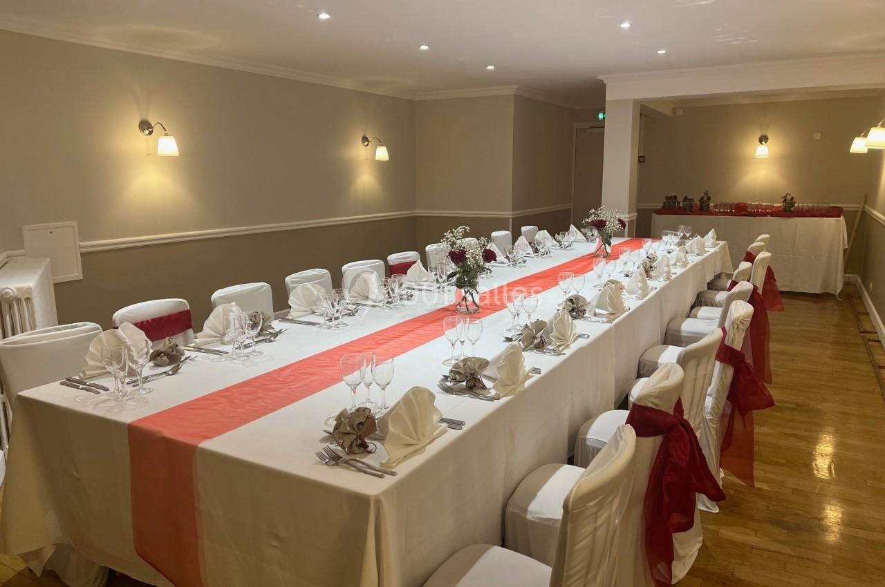 Grande table rectangulaire dressée pour un repas, avec nappes blanches, chemins de table rouges et chaises décorées.