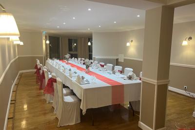Salle lumineuse avec une grande table rectangulaire dressée, entourée de chaises décorées, parquet clair et fenêtres donnant…