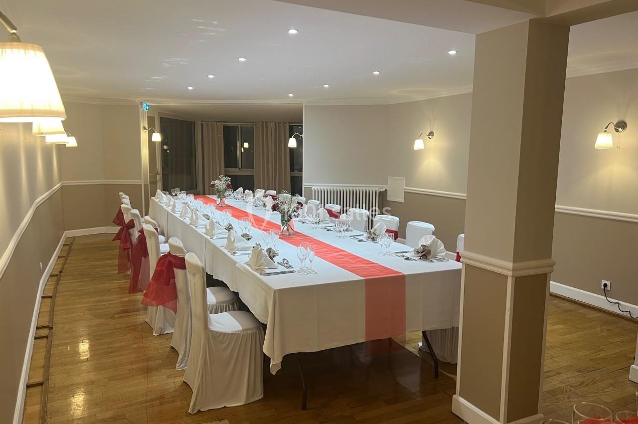 Salle de réception avec une grande table dressée, nappes blanches, chemin de table rouge et chaises décorées.