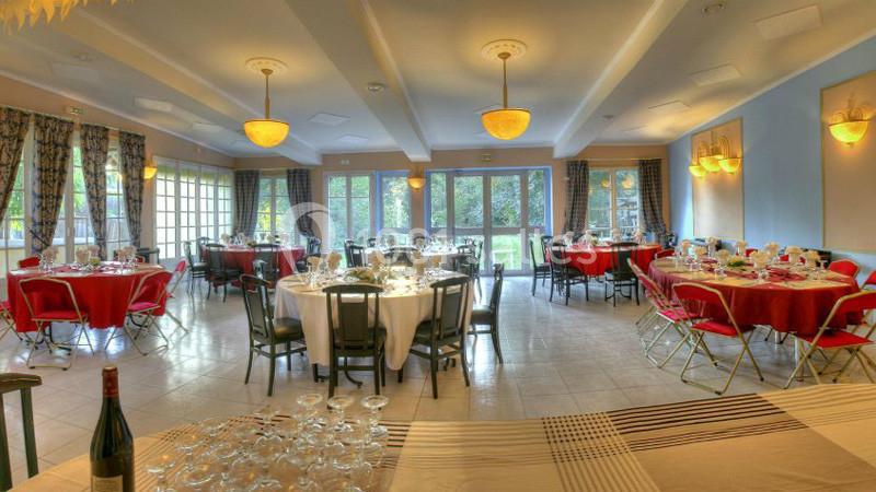 Salle de réception lumineuse avec tables rondes dressées, nappes blanches et rouges, et vue sur un jardin.