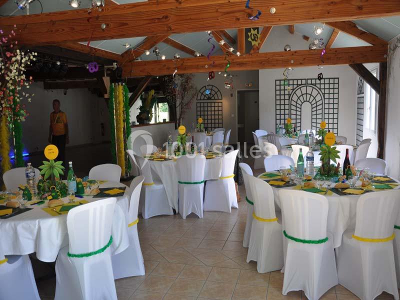 Salle décorée pour un événement, avec des tables rondes dressées, nappes blanches et ornements aux couleurs vives.