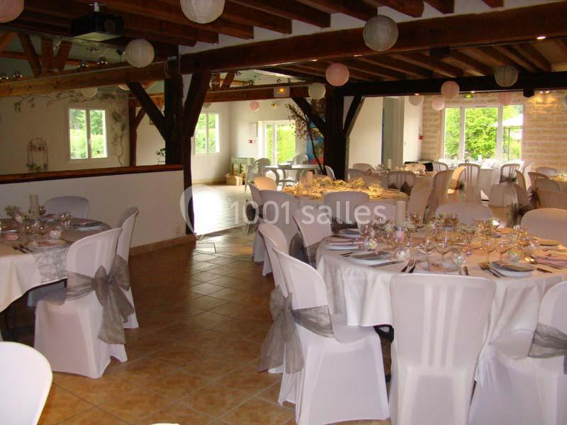 Salle de réception décorée avec des tables rondes dressées, chaises blanches et poutres apparentes en bois.