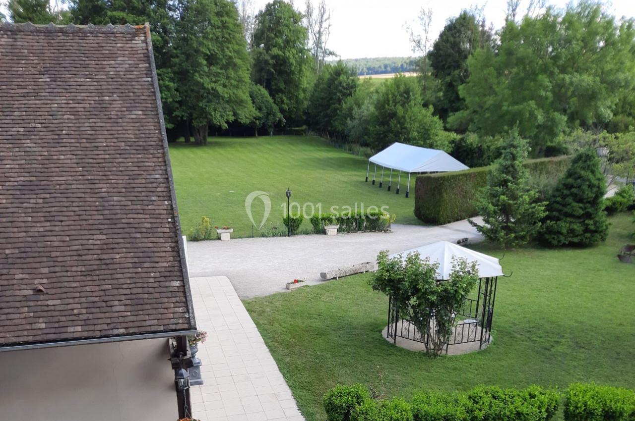 Vue d'un jardin spacieux avec pelouse, arbres, tonnelle et barnums, bordé par une maison et une terrasse en premier plan.