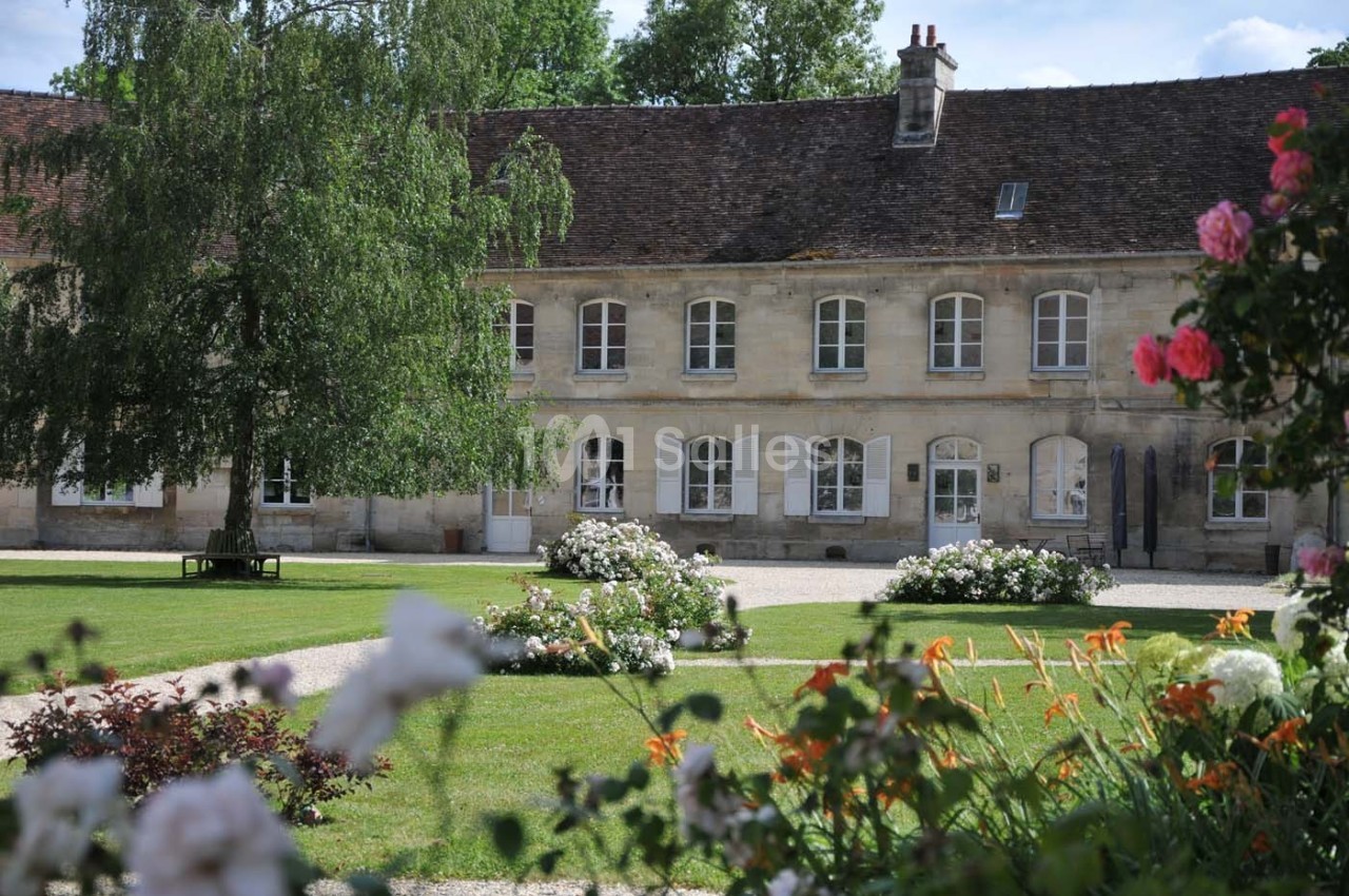 Bâtiment en pierre avec toit en tuiles, entouré d'un jardin fleuri et d'arbres par une journée ensoleillée.