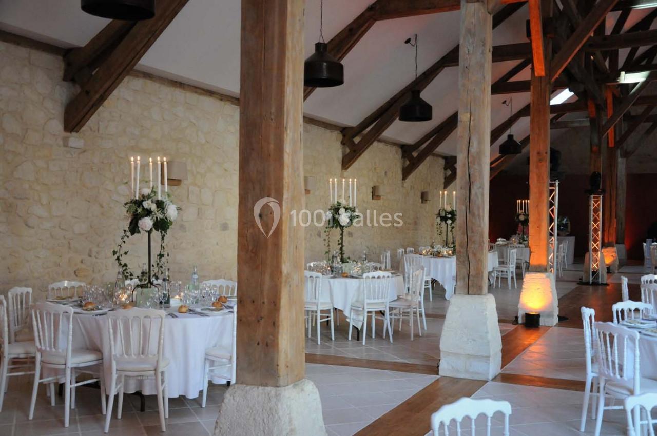 Salle de réception avec tables rondes décorées, chandeliers et murs en pierre apparente sous une charpente en bois.