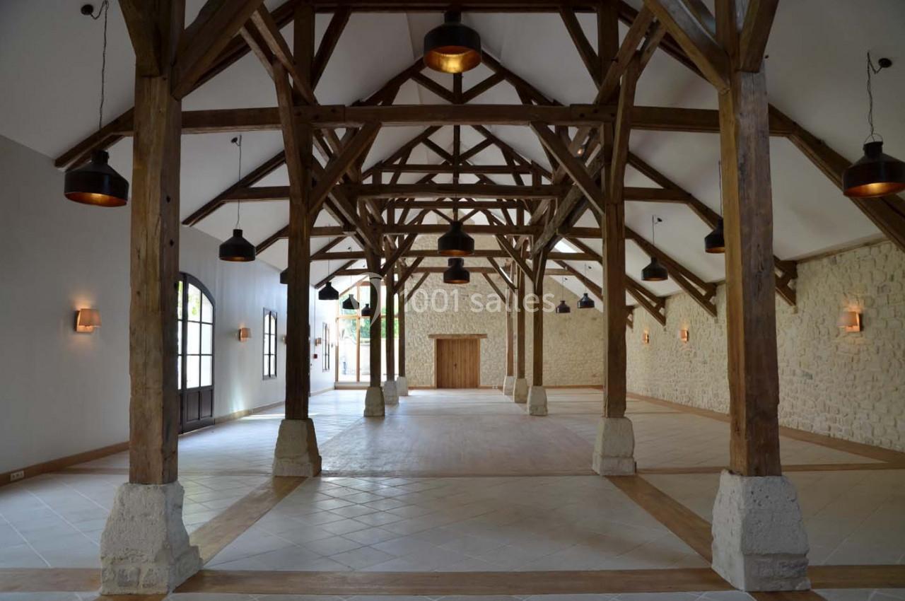 Grande salle avec charpente en bois apparente, murs en pierre claire, sol carrelé et grandes ouvertures vitrées.