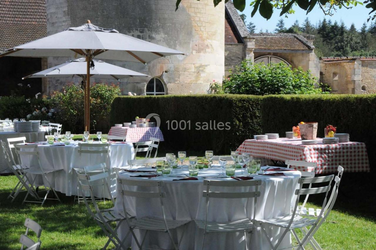 Tables rondes dressées avec nappes blanches et chaises pliantes dans un jardin, sous des parasols et près de haies taillées.