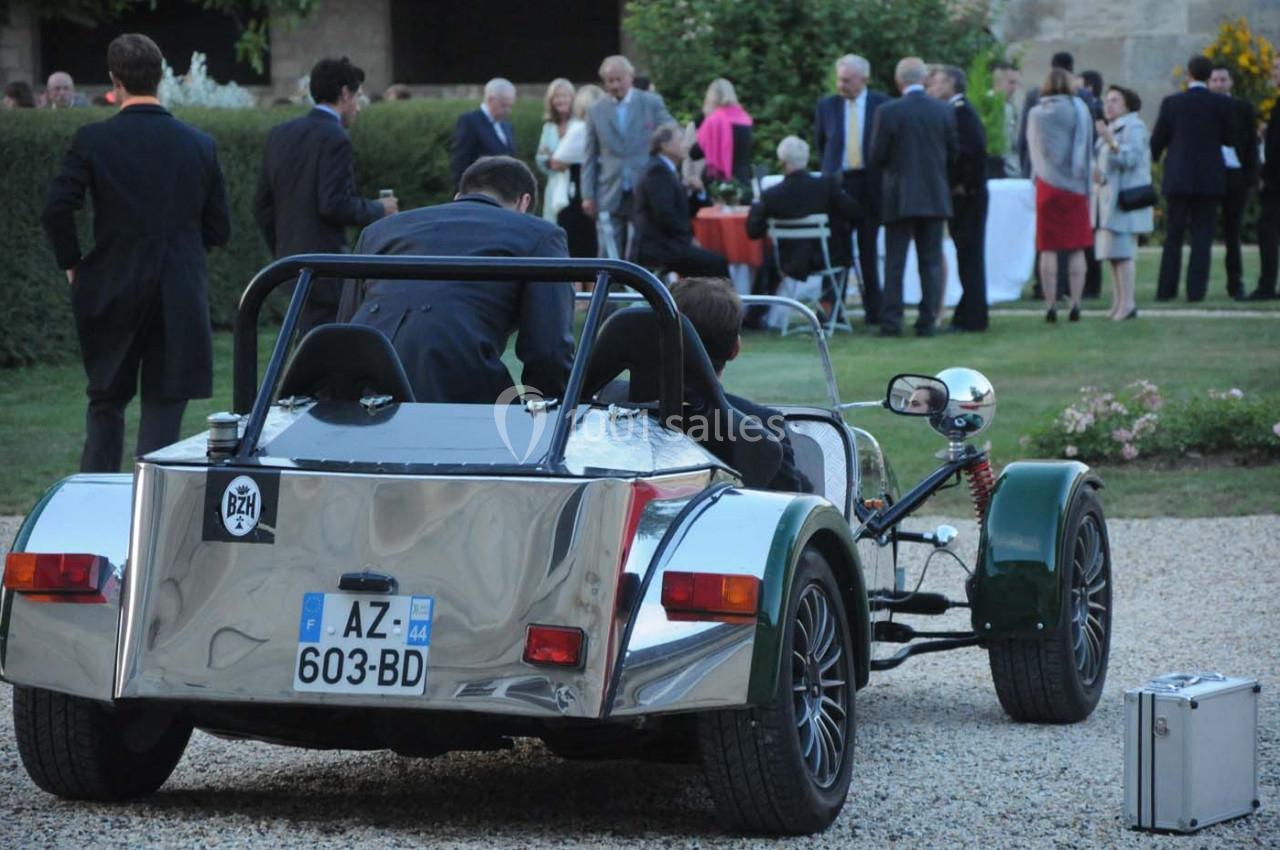 Deux hommes assis dans une voiture de sport rétro stationnée sur une allée gravillonnée lors d'un événement en plein air.