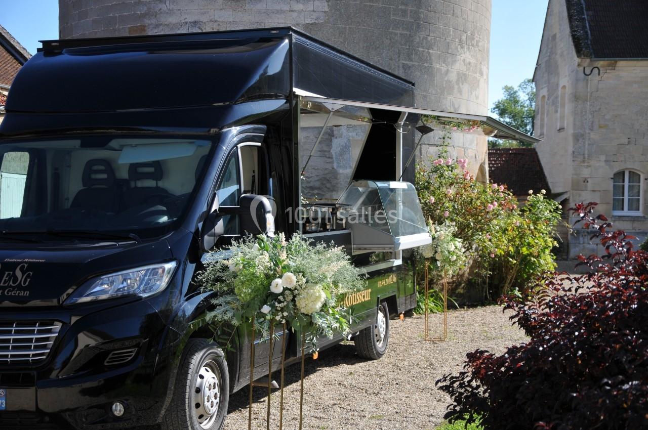 Camion noir aménagé en food truck, décoré de fleurs blanches, stationné devant un bâtiment en pierre.