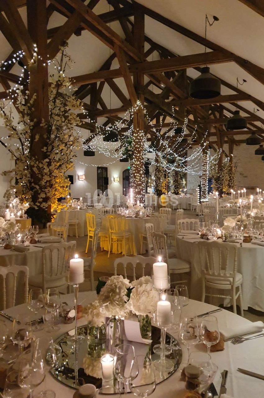 Salle de réception décorée pour un mariage, avec tables dressées, bougies, guirlandes lumineuses et arbres fleuris.