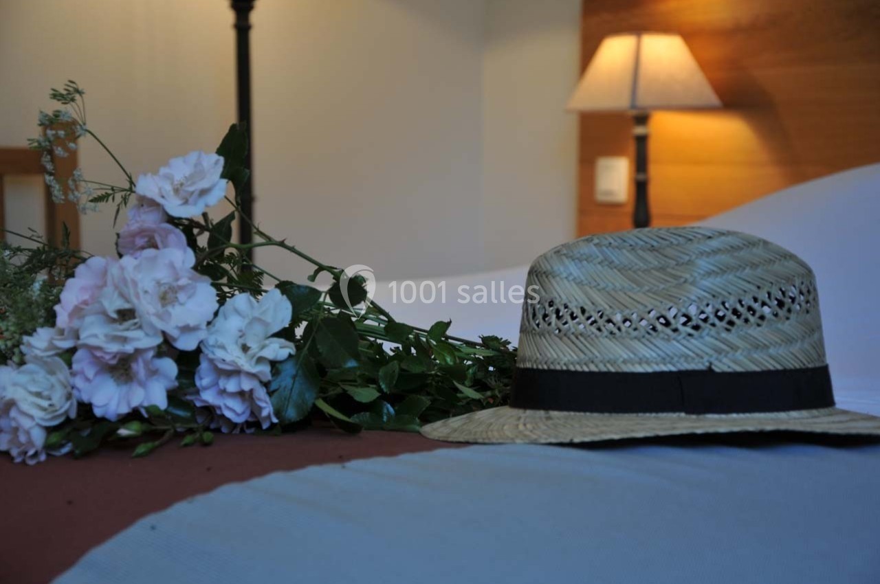 Chapeau de paille posé sur un lit avec un bouquet de fleurs roses et une lampe allumée en arrière-plan.