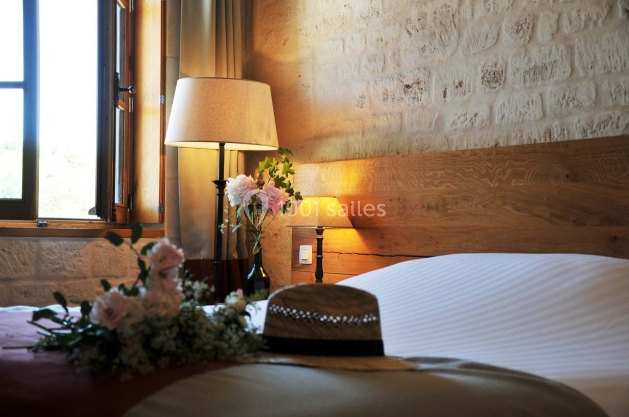 Lit avec draps blancs dans une chambre en pierre, éclairée par une lampe et une fenêtre ouverte, décorée de fleurs et d'un…